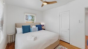 Un dormitorio con una cama, ventilador de techo, ventana, puerta y una mesita.