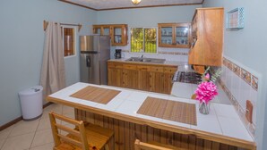 A kitchen with wooden cabinets, a white countertop, and a dining area with two wooden chairs. - Roatan Ocean View - Ground Floor 2 Bedroom Apts (Roatan)