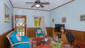 A living room with a wooden ceiling fan, a television on a wooden stand, a blue sofa, a wooden dining table with a potted plant, and a window with a curtain. - Roatan Ocean View - Ground Floor 2 Bedroom Apts (Roatan)