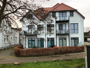 Um edifício residencial com vários andares, telhado com telhas, janelas amplas e um jardim ajardinado.