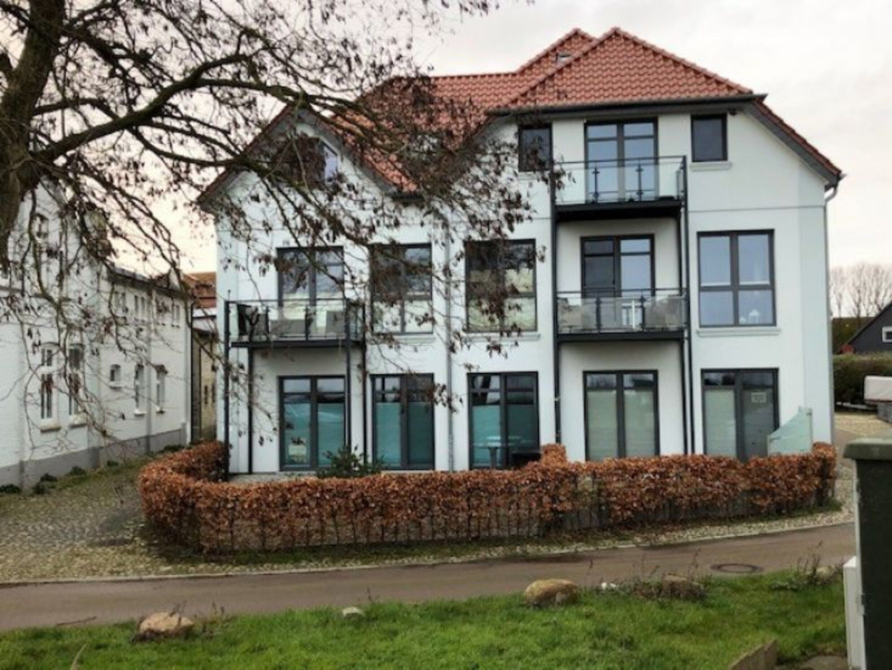 Ein mehrstöckiges Wohnhaus mit Ziegeldach, großen Fenstern und einer gepflegten Hecke davor.