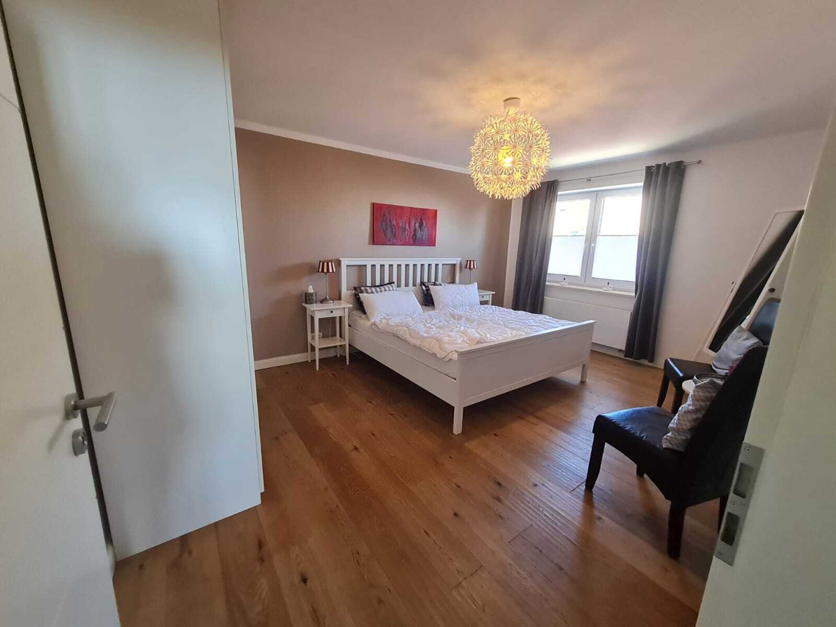 Ein Schlafzimmer mit einem weißen Bett, Holzboden, einem Nachttisch mit Lampe, einer Deckenleuchte und einem Fenster mit Vorhängen.