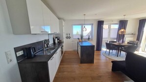 Moderni keittiö, jossa on valkoiset kaapit, keskellä oleva keittiösaareke sekä ruokailutila, jossa on tummanvärinen sohva ja pöytä.