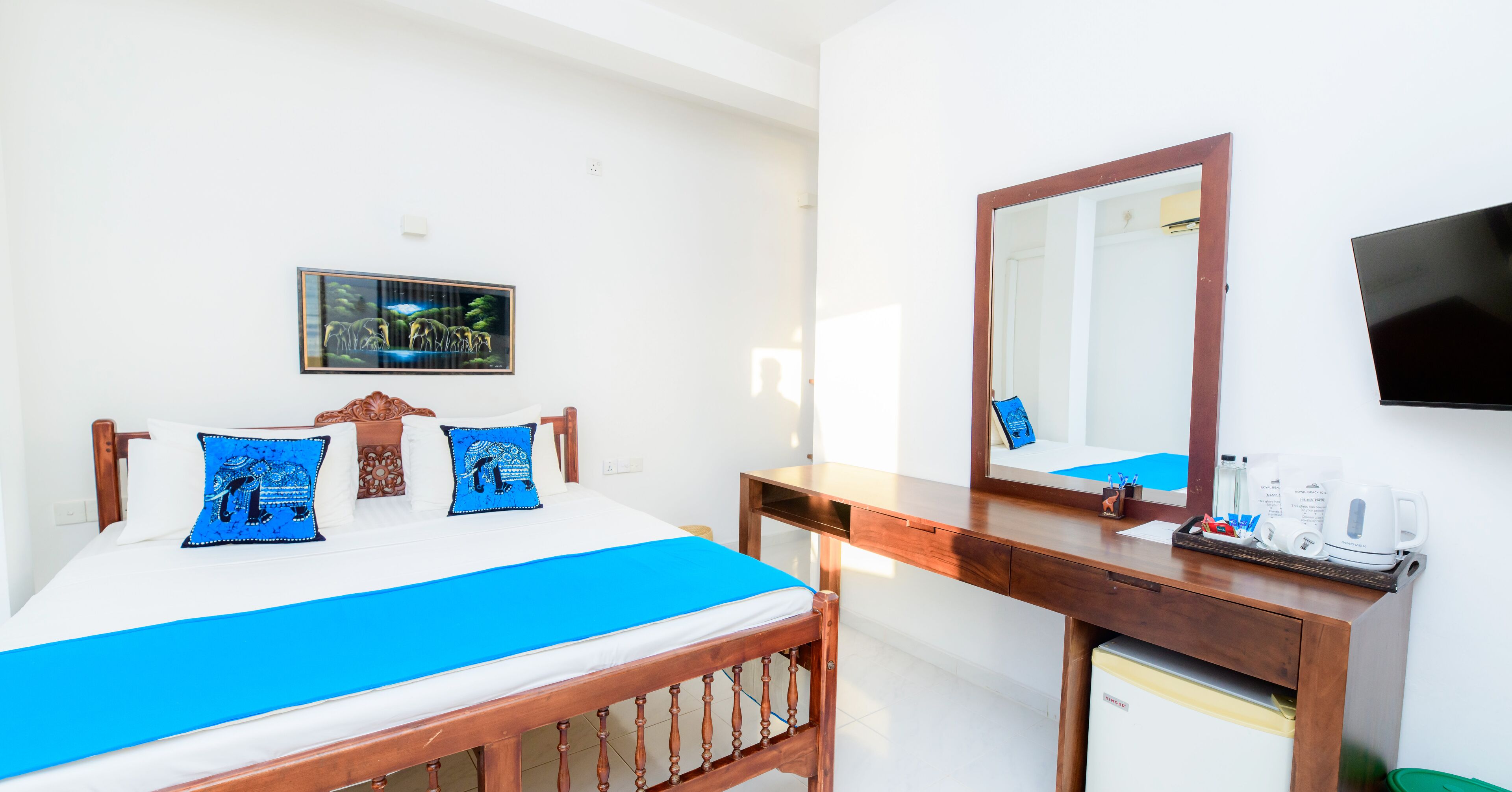 Habitación de hotel con cama, escritorio de madera, espejo, televisor y refrigerador pequeño.