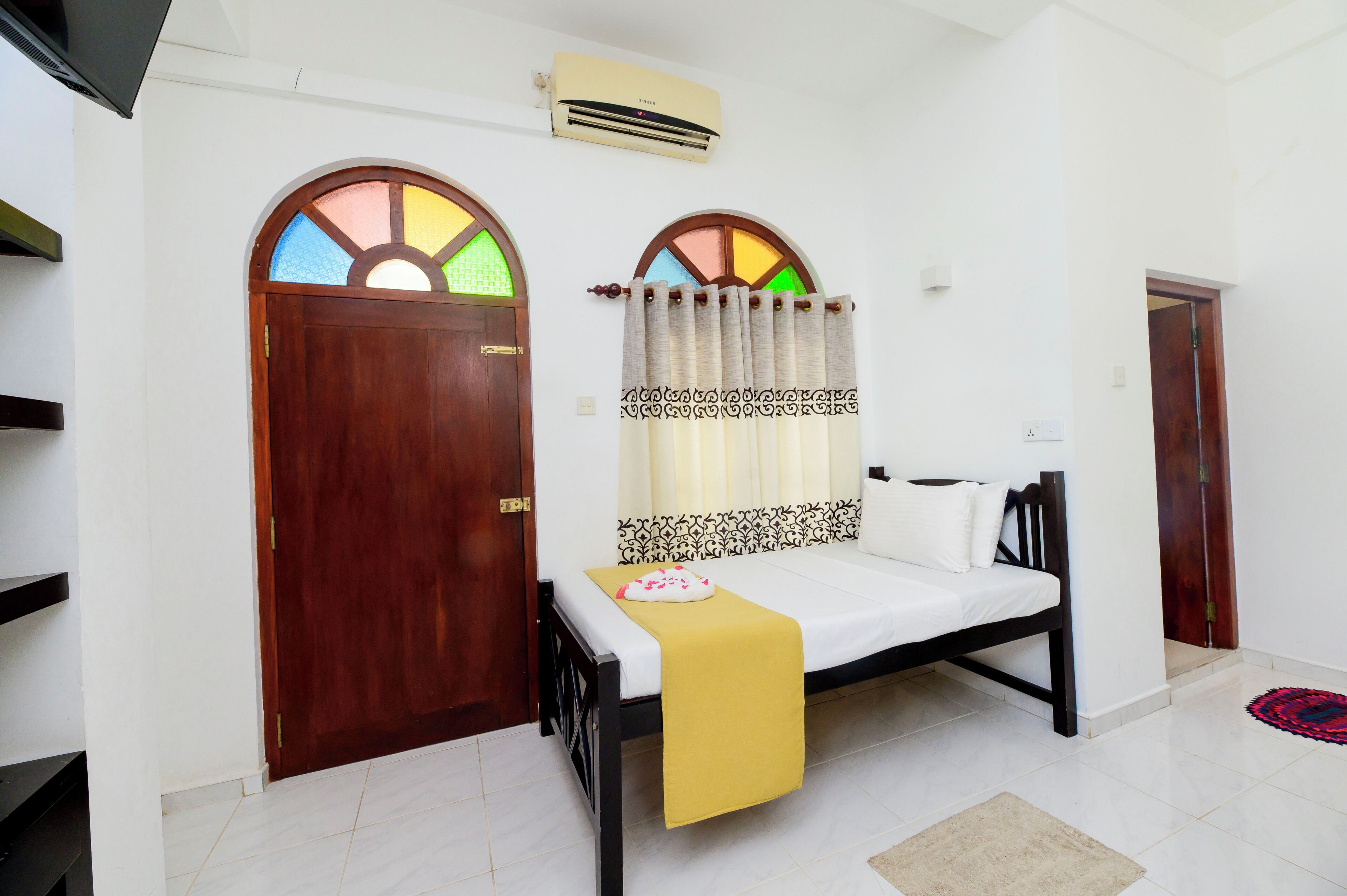 A room with a bed, a wooden door, an air conditioning unit, and a window with a stained glass design.