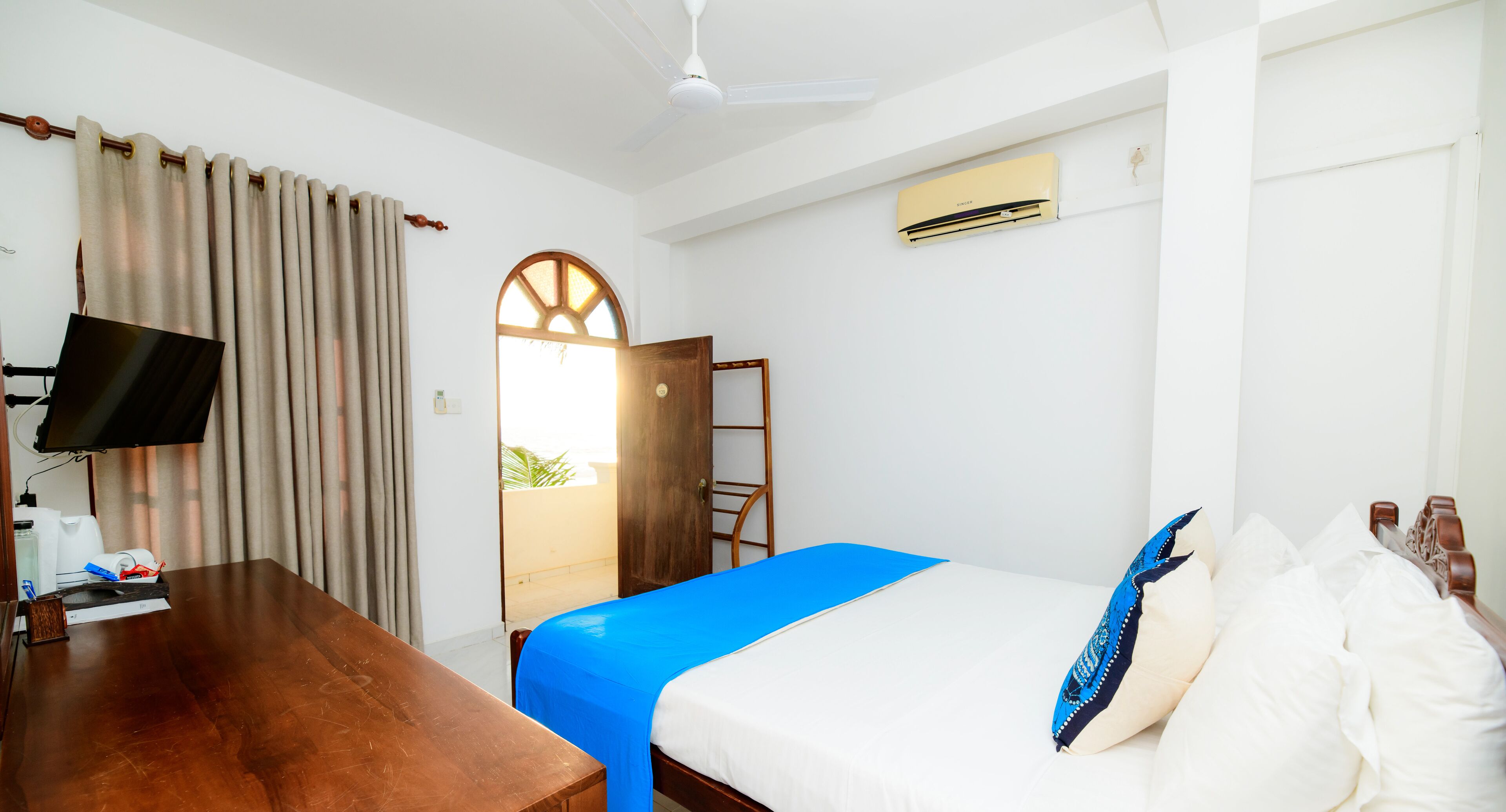 Quarto de hotel com uma cama, uma escrivaninha, televisão, janela com cortinas e ar condicionado.