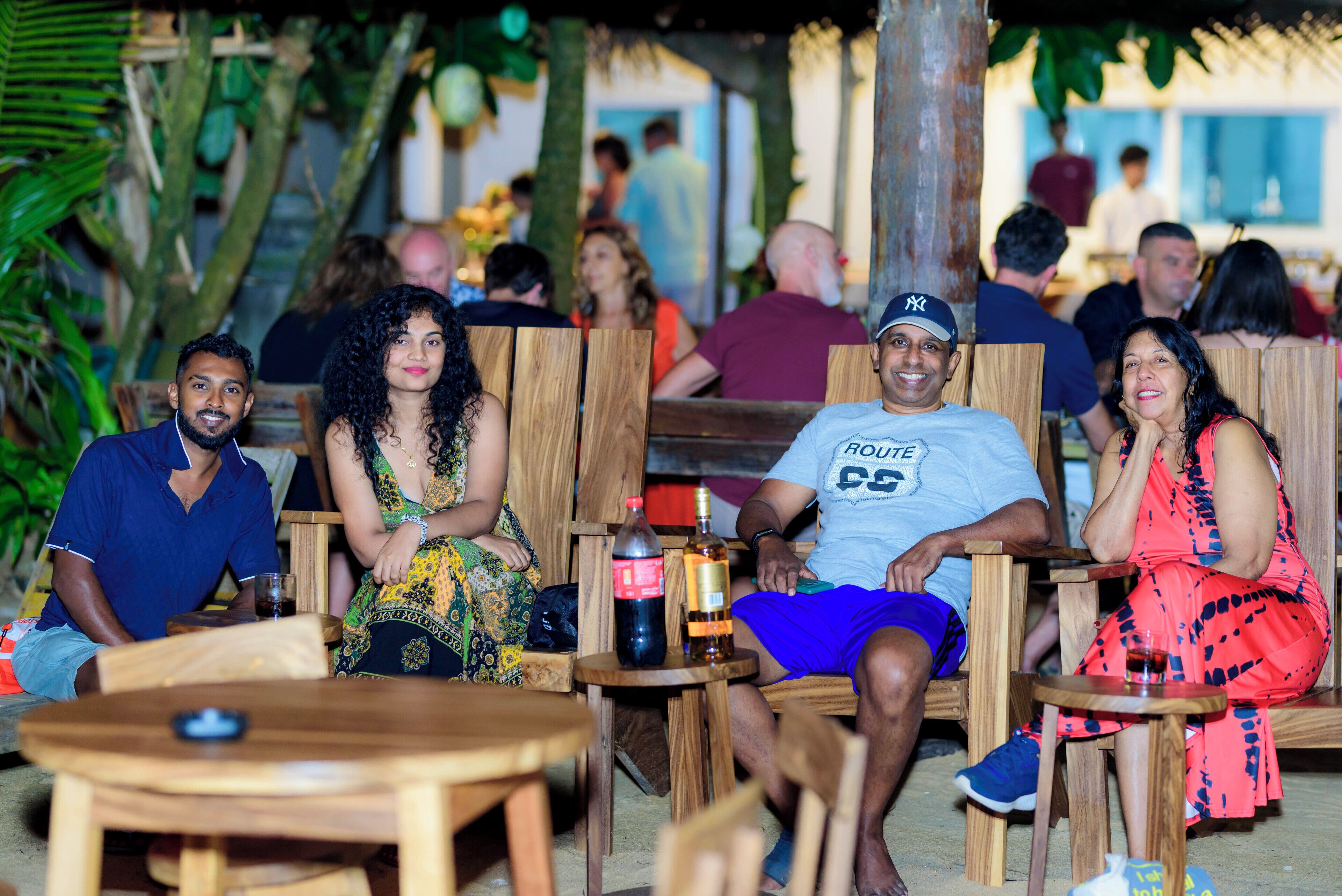 A group of people seated at wooden tables in an outdoor setting with tropical plants in the background.