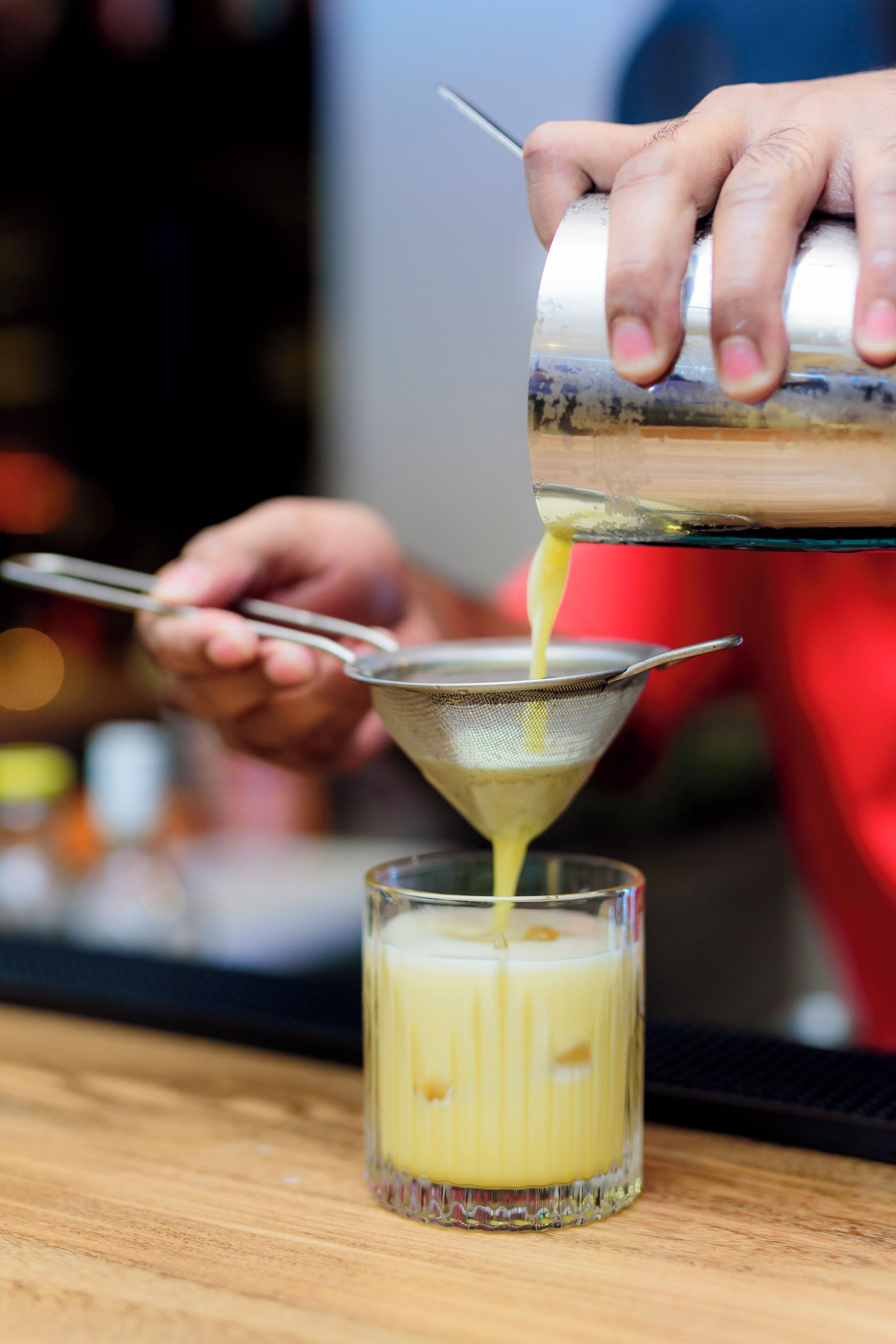 A person pouring a yellow liquid from a shaker into a glass through a strainer.