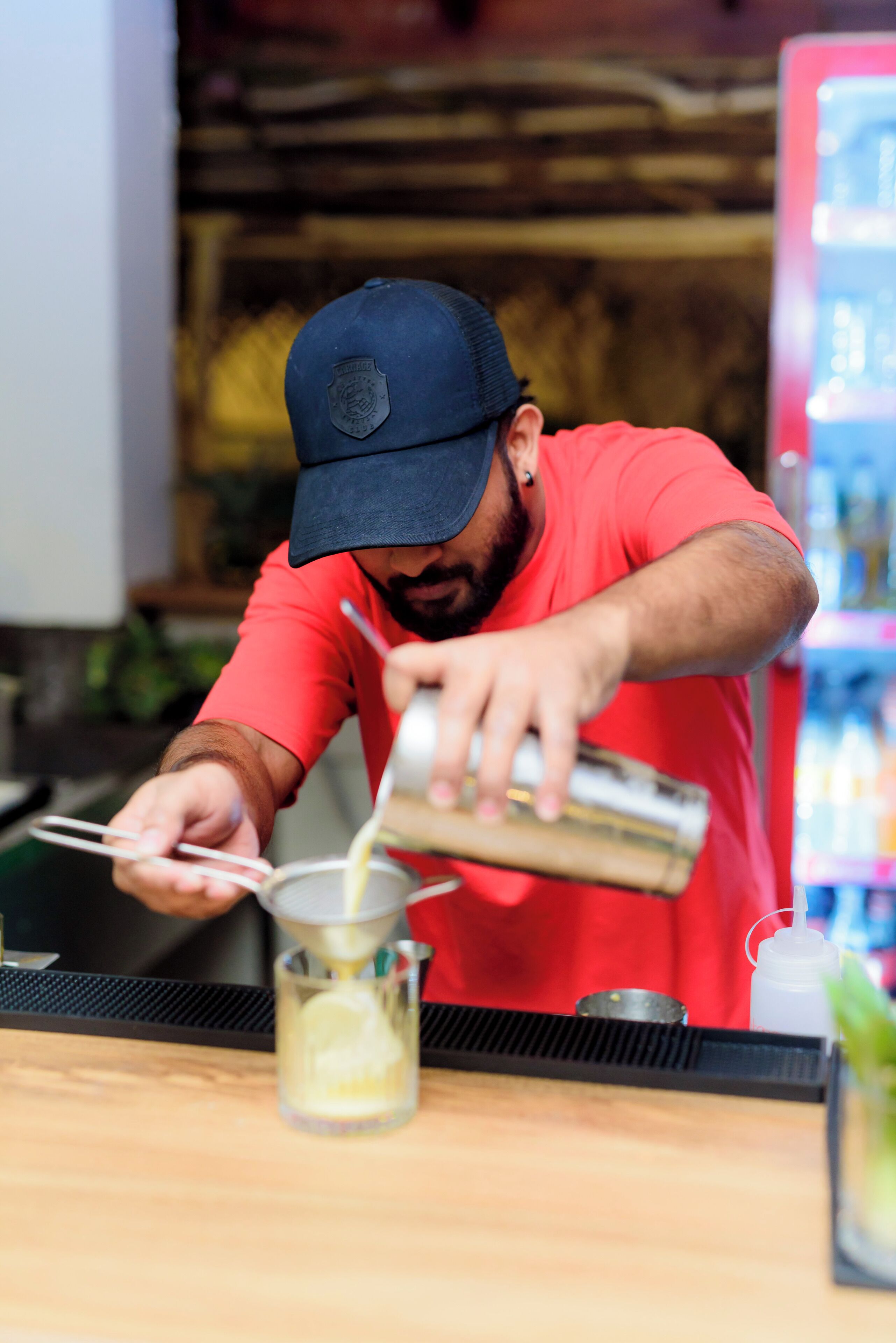 A person preparing a drink at a bar with a counter and a refrigerator in the background.