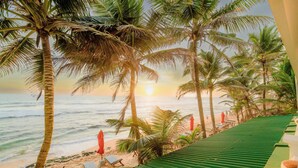 Vista de playa con palmeras, un atardecer y una playa con sillas y sombrillas.