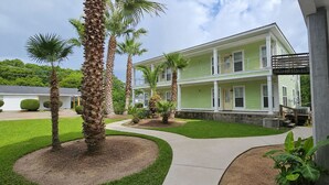 A two-story building with a green facade, a covered porch, and a landscaped garden with palm trees. - Ultimate Group Stay · 2 Full Apts · 6BR · Parking (Moss Point)
