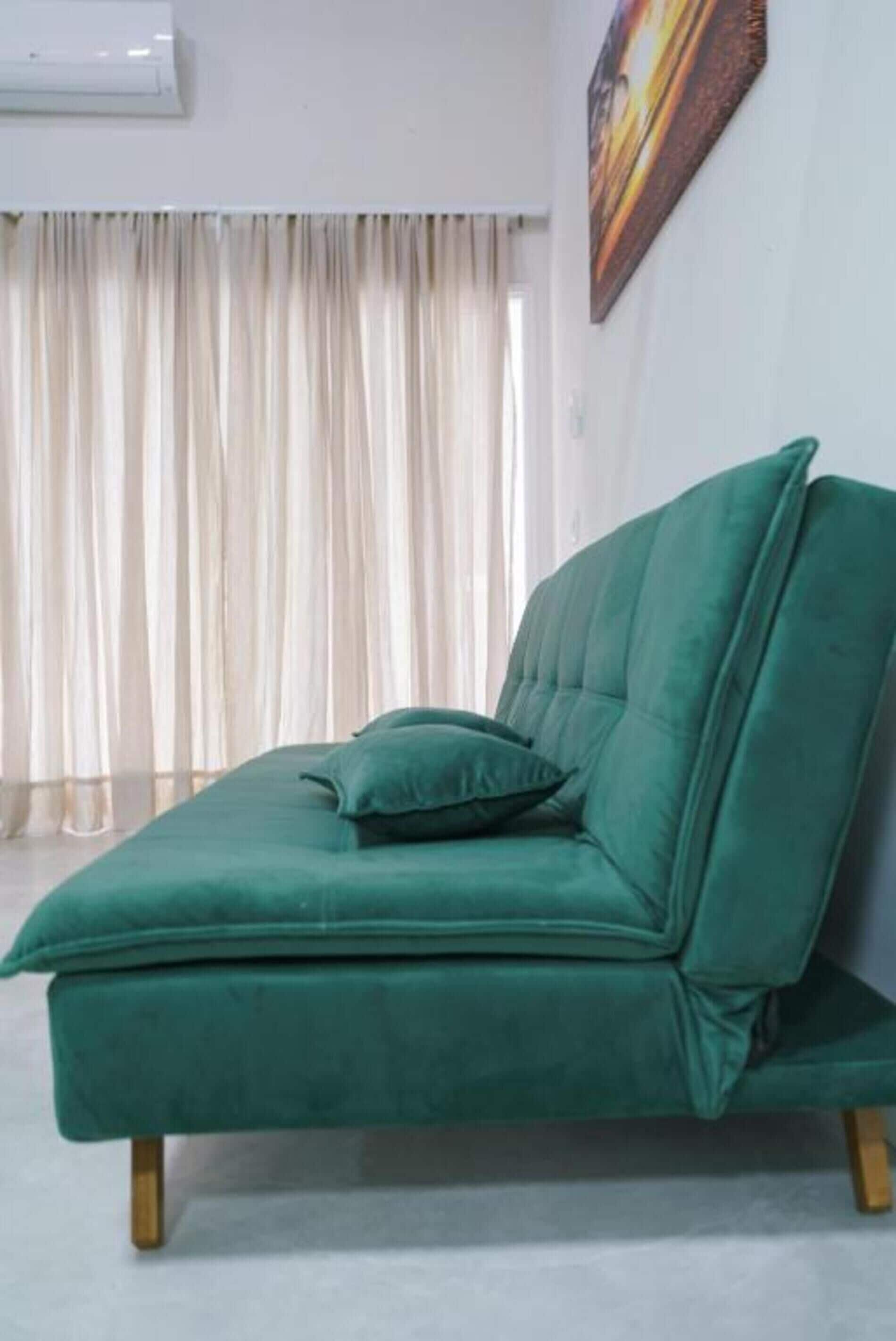 A green sofa with wooden legs in a room with a white air conditioner and a painting on the wall.