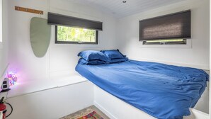 Un dormitorio con una cama individual, dos ventanas con persianas, un tapete pequeño y un estante fijado a la pared.