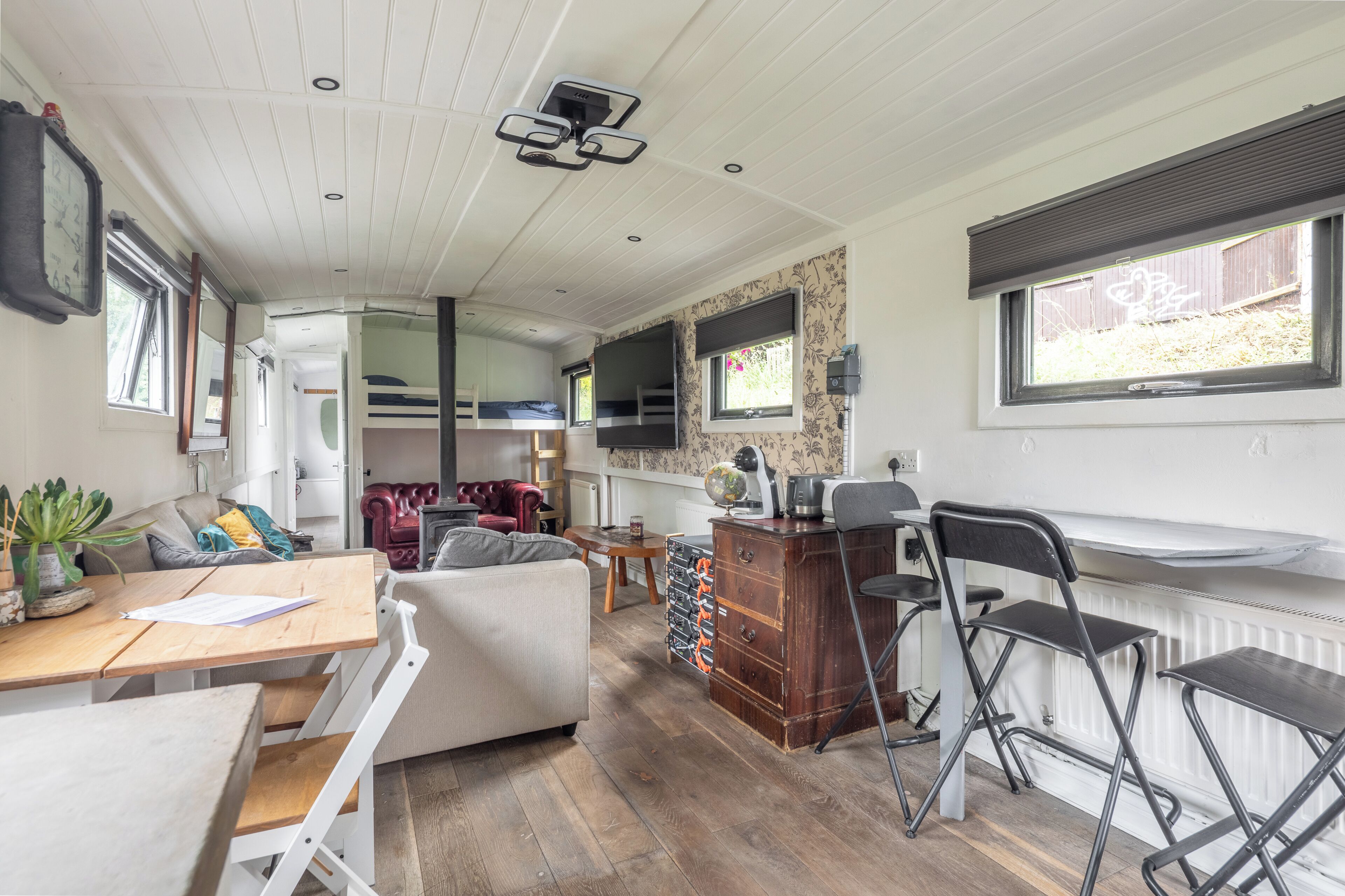 Una habitación compacta y bien iluminada, con piso de madera, sofá blanco, mesa de comedor con sillas, pequeña área de cocina y ventana con persianas.