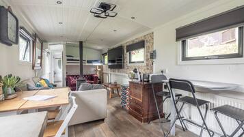 Una habitación compacta y bien iluminada, con piso de madera, sofá blanco, mesa de comedor con sillas, pequeña área de cocina y ventana con persianas.