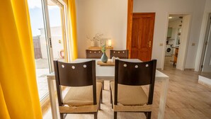 Uma área de refeições com mesa branca e quatro cadeiras, um vaso com flores e uma porta de madeira ao fundo.