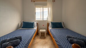 Quarto pequeno e limpo com duas camas de solteiro, uma mesinha com um vaso, e uma janela com cortinas.