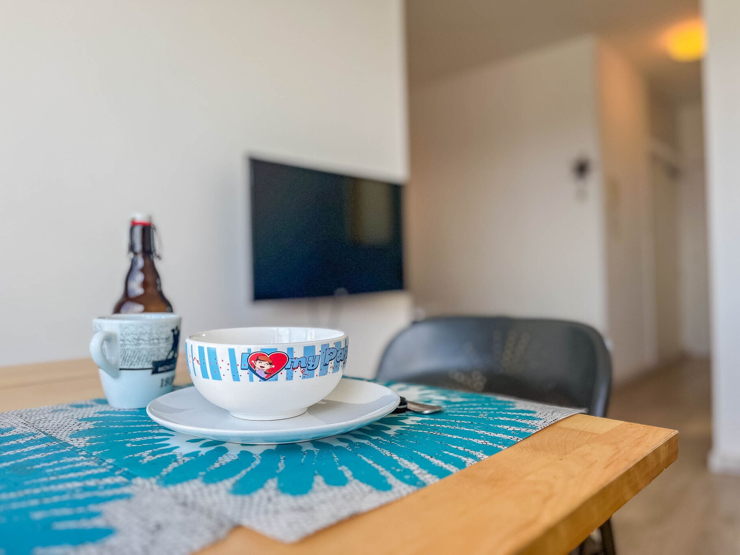 Een eettafel met een kom en een fles, een stoel en een televisie op de achtergrond.