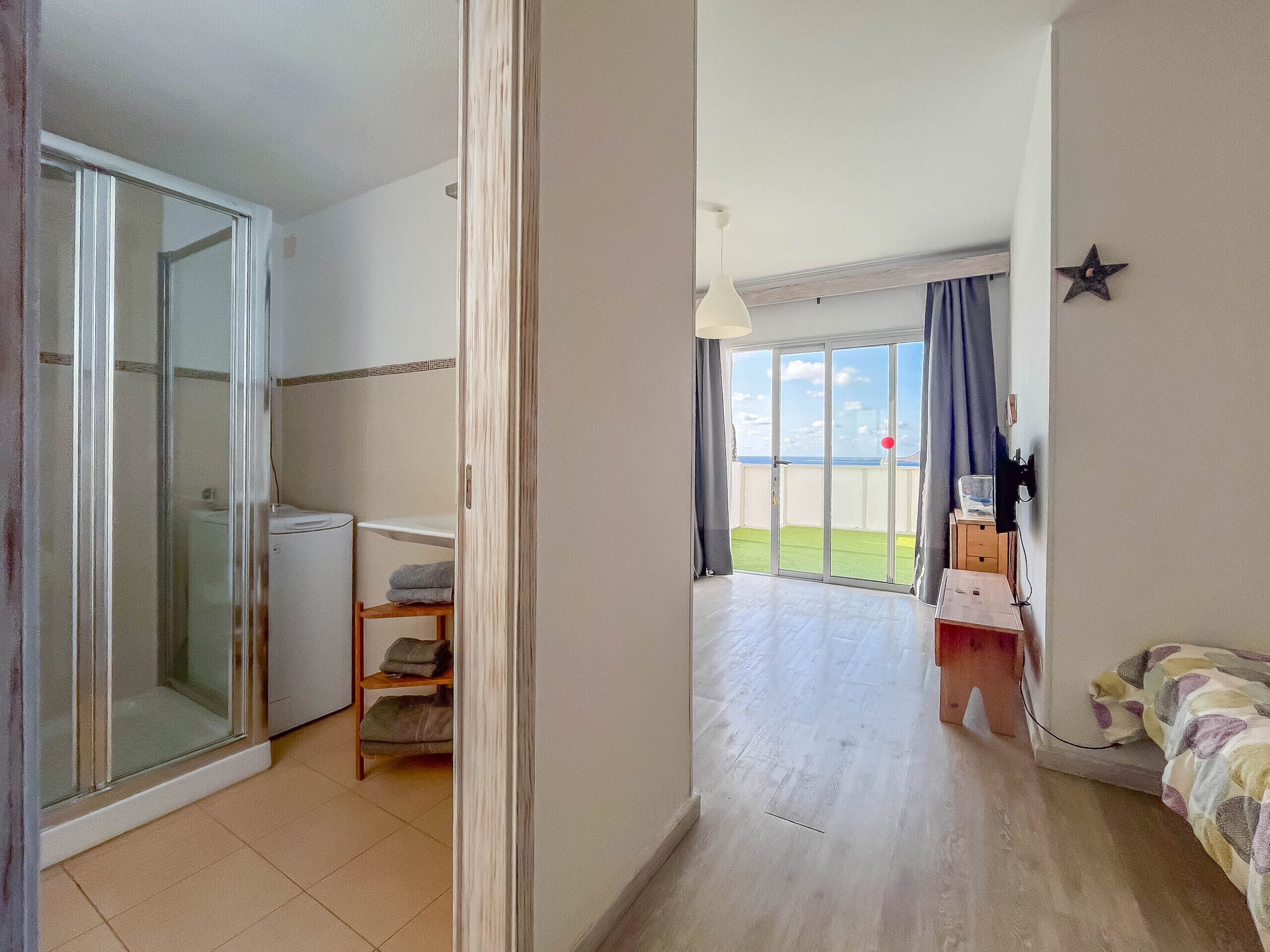 A hotel room with a sliding glass door leading to a balcony, a small kitchenette, a bed with a colorful blanket, and a star decoration on the wall.