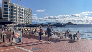 Een restaurant aan het water met terras, een bord waarop 'La Paella' staat en een modern gebouw op de achtergrond.