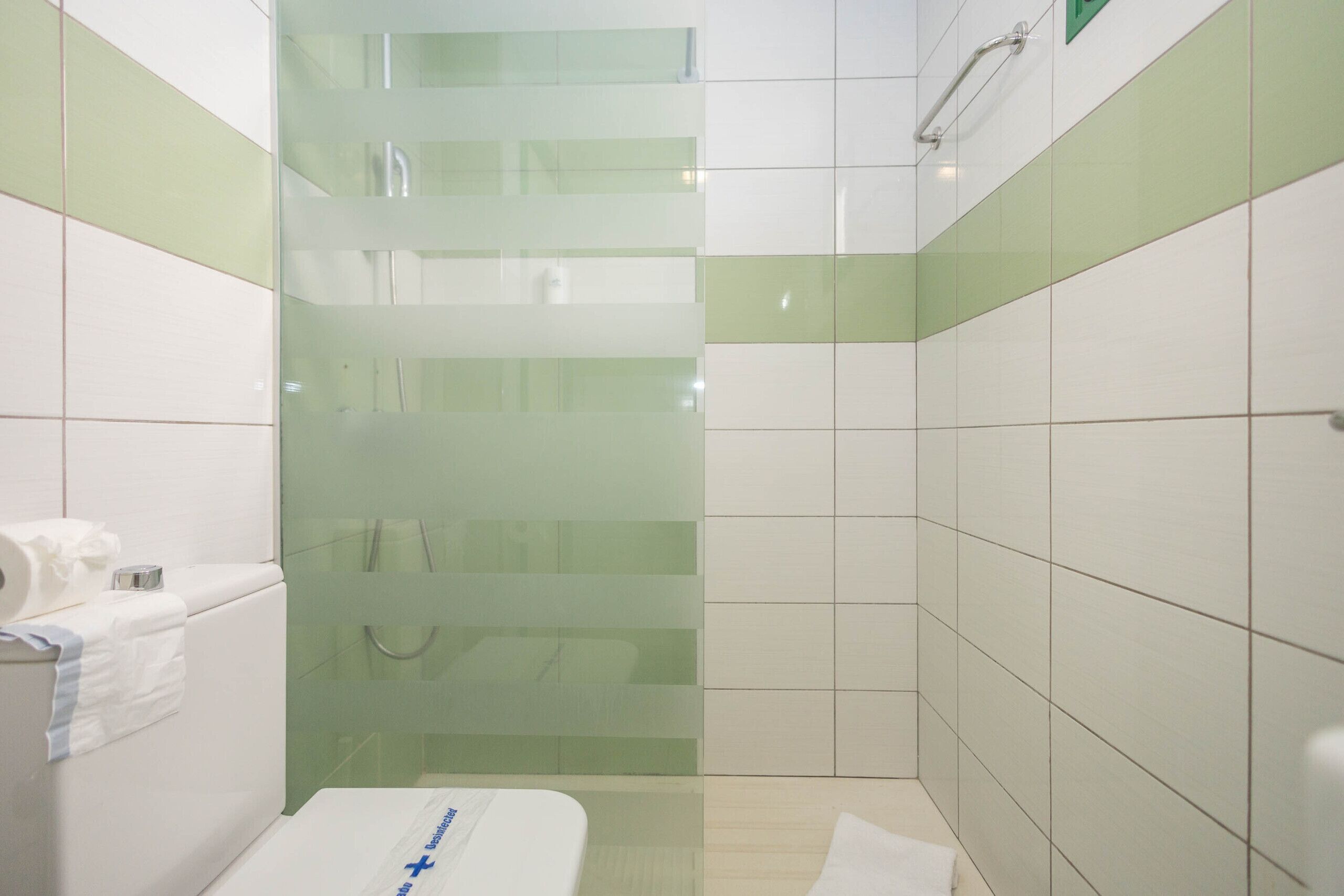A bathroom with a glass shower enclosure, white and green tiled walls, a white sink, and a white toilet.