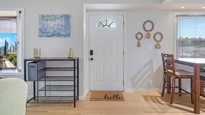 A modern living room with a white door, a painting, a shelf with decorative items, a wooden dining table, and a chair. - The Calm & Comfy Nest (Minneapolis)