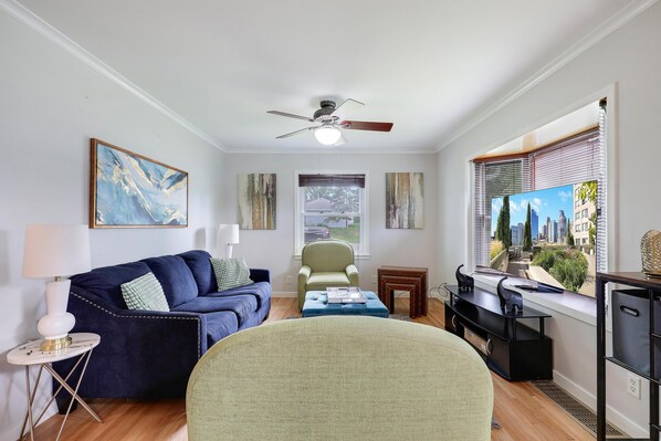 A living room with a dark blue sofa, a green armchair, a small table with a lamp, and a cityscape view through the window. - The Calm & Comfy Nest (Minneapolis)