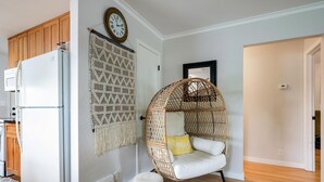 A kitchen with a white refrigerator, wooden cabinets, a macrame wall hanging, a wicker chair with a cushion, and a small ottoman. - The Calm & Comfy Nest (Minneapolis)