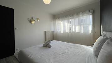 Una cama bien tendida con una toalla enrollada, una mesita de noche, una lámpara de pared y una ventana con cortinas traslúcidas.
