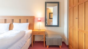 Habitación de hotel con cama, mesita de noche con lámpara, un pequeño banco, un espejo y armarios de madera.