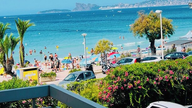 A beach with people swimming and sunbathing, cars parked along the shore, and a clear view of the sea and distant land. - Seafront apartment with sea view. Everything within walking distance with parking lot. (Saint-Cyr-sur-Mer)