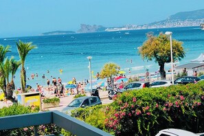 Una spiaggia con persone che nuotano e si prendono il sole, auto parcheggiate lungo la riva e una vista chiara del mare e della costa in lontananza.