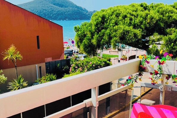 A balcony with a view of the sea, a building with a red facade, and a tree.