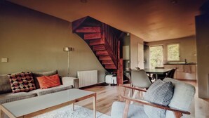 Ein modernes Wohnzimmer mit einer Holztreppe, einem Sofa mit gemusterten Kissen, einem Couchtisch und einem Essbereich mit Stühlen.