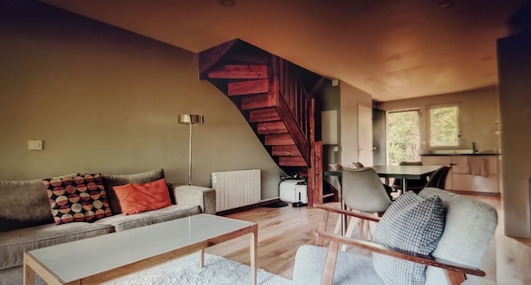 A modern living room with a wooden staircase, a sofa with patterned cushions, a coffee table, and a dining area with chairs.