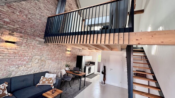 A loft-style living area with exposed brick walls, a wooden staircase, and a mezzanine with a black railing.