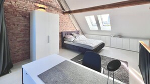 A bedroom with a loft-style design, featuring exposed brick walls, a bed with a headboard, a desk, and a chair. There are skylights allowing natural light into the room.