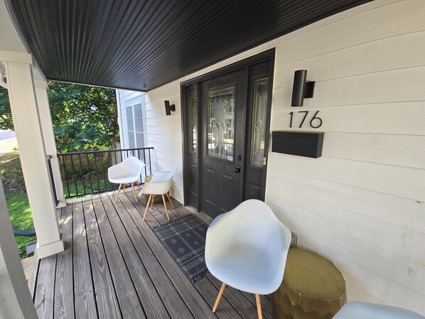 A wooden deck with two white chairs and a small round table, a black door with a number plaque, and a white building with a dark roof.