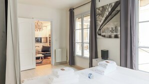 Una habitación de hotel con cama, radiador, ventana con cortinas, un mural de paisaje urbano y una puerta que da a otra habitación.