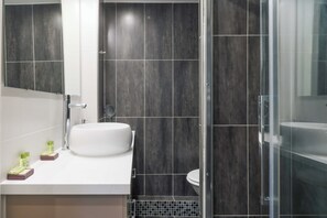 A modern bathroom with a white sink, dark tiled walls, and a glass shower enclosure. - Comfortable Apartment - 1br/2p - Arts et Metiers (Paris)