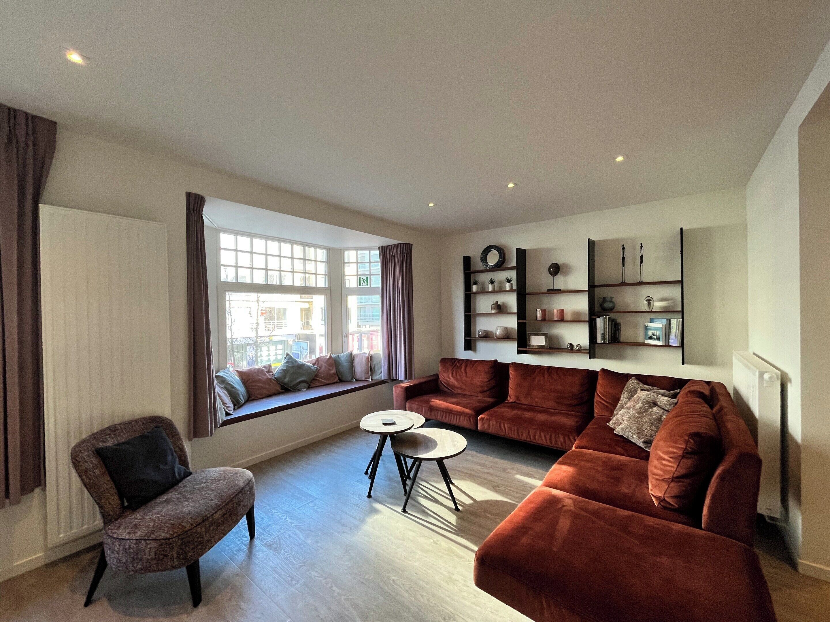 A modern living room with a sofa, armchair, and a small table. There is a window with curtains and a built-in bench. Shelves with decorative items are against the wall.