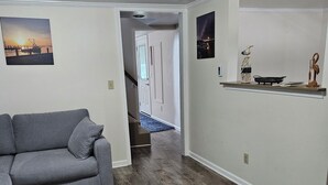A living room with a gray sectional sofa, wooden flooring, a hallway leading to a door, and a shelf with decorative items.