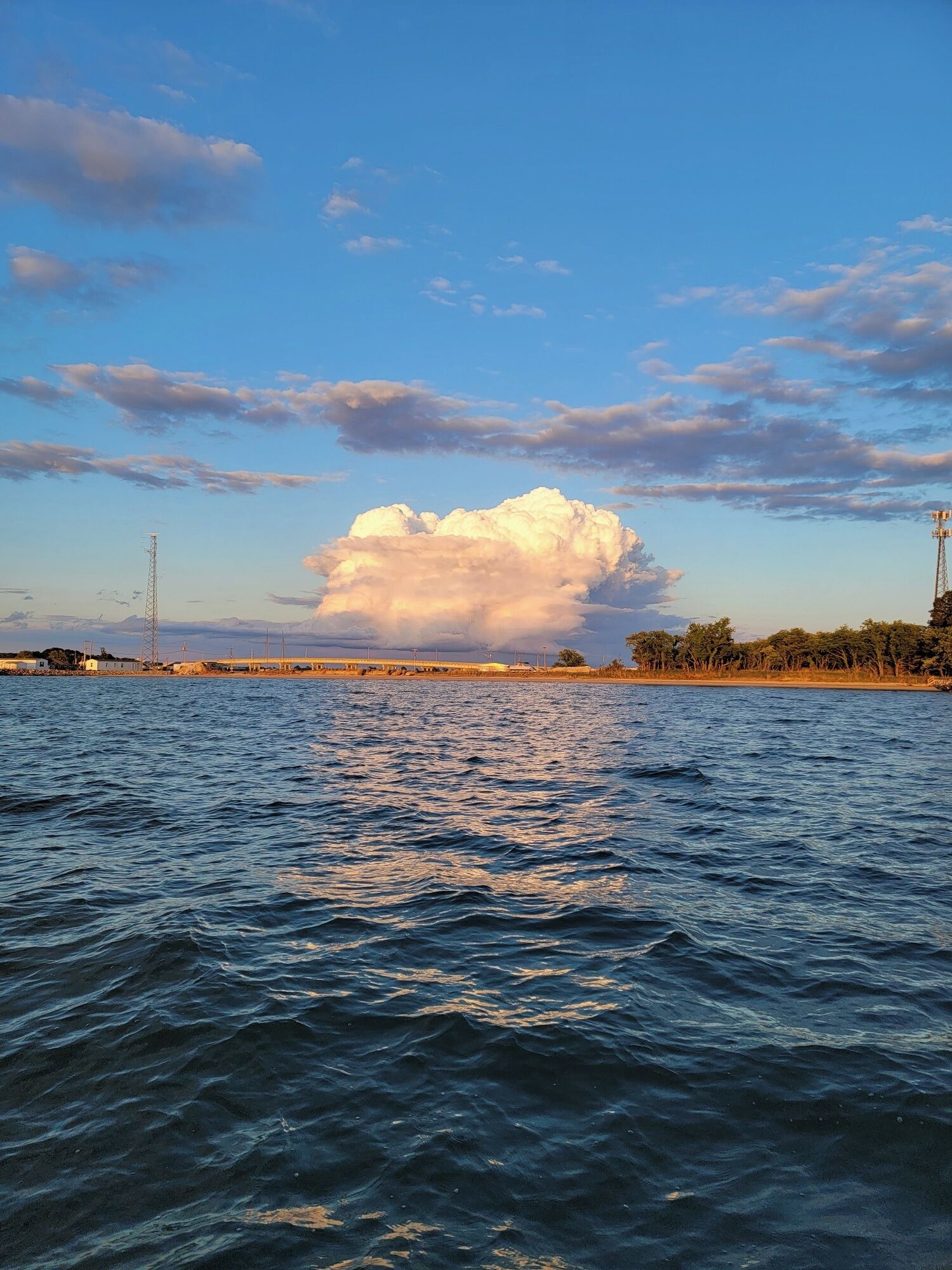 Blick auf die Küste mit einer großen Wolkenformation über dem Wasser, einer Brücke im Hintergrund und Stromleitungen.