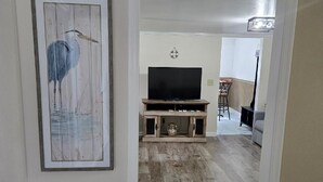 A hallway with a framed artwork of a bird, a wooden entertainment unit with a TV, and a dining area with a bar stool.