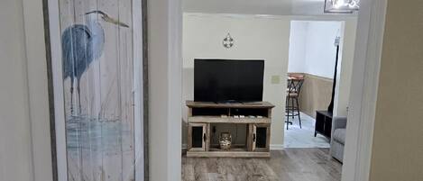 Een gang met een lijstwerk waarop een vogel afgebeeld is, een houten entertainmentkast met een televisie, en een eethoek met een barkruk.