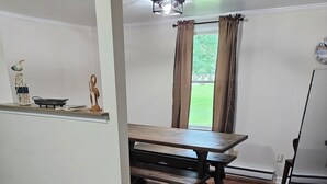 A dining area with a wooden table and benches, a small kitchen counter with decorative items, and a view through a window with curtains.