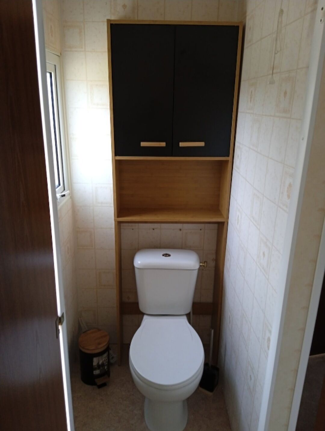 Une petite salle de bain avec des toilettes blanches, un meuble en bois au-dessus et une petite poubelle sur la gauche.