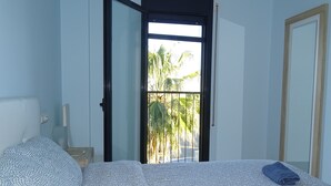 A bedroom with a bed, a nightstand, a lamp, and a view of palm trees through an open door.