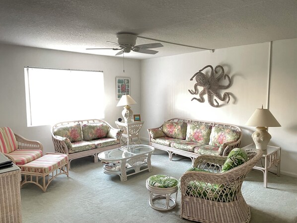 A living room with wicker furniture, a ceiling fan, and a large window. - Beach Bliss Awaits: Immaculate Condo Just Steps from Your Private Beach Oasis! (Key Colony Beach)