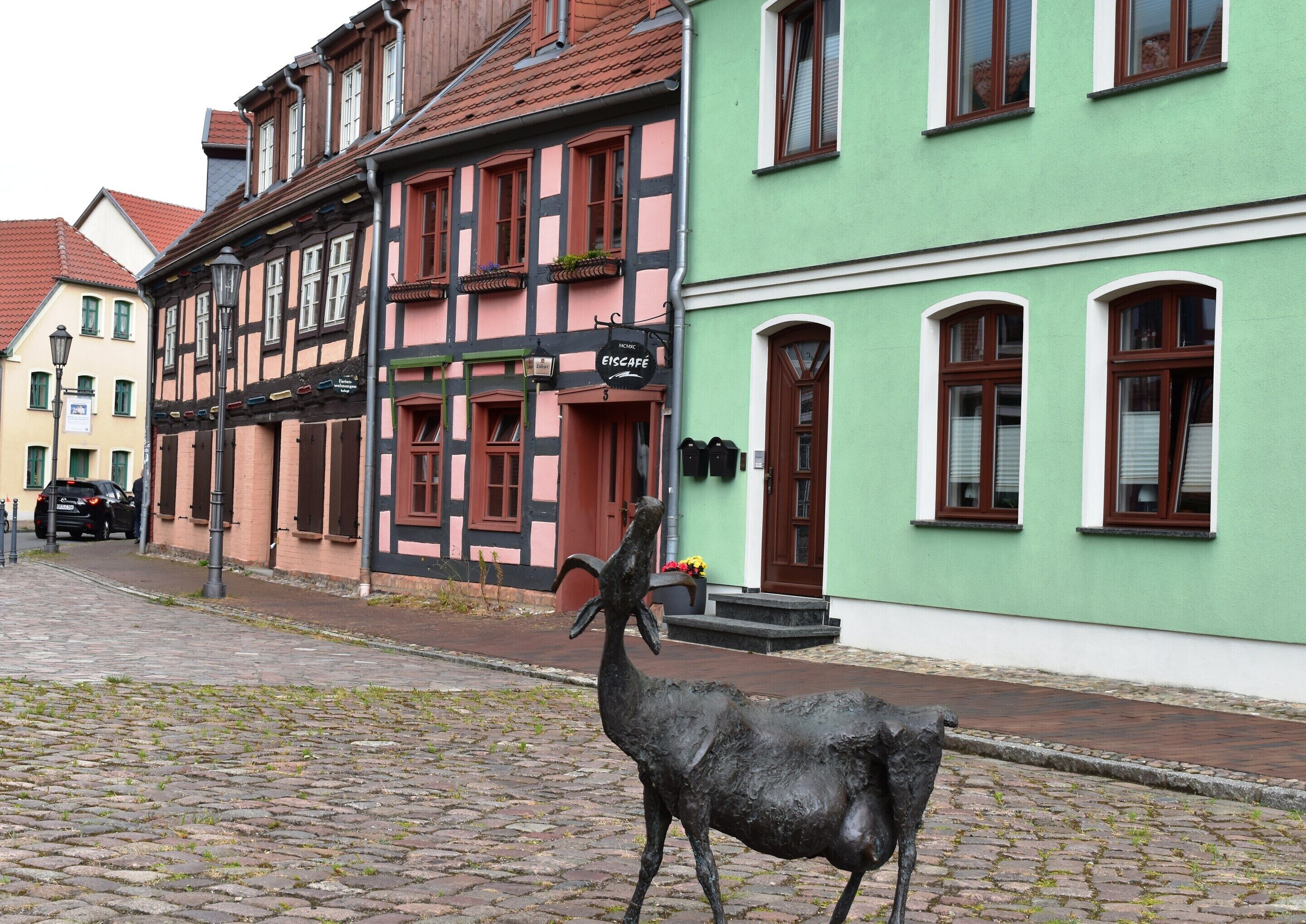 Eine Kopfsteinpflasterstraße mit einem grünen Gebäude mit traditioneller Fassade, einer schwarzen Metallskulptur und einem geparkten Auto.